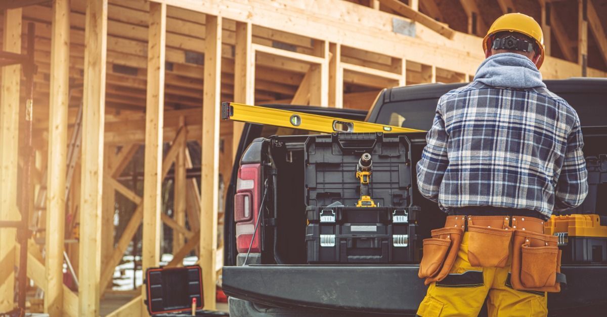 A construction worker wearing a toolbelt behind a pickup truck full of thick, black crates. The truck is on a jobsite.