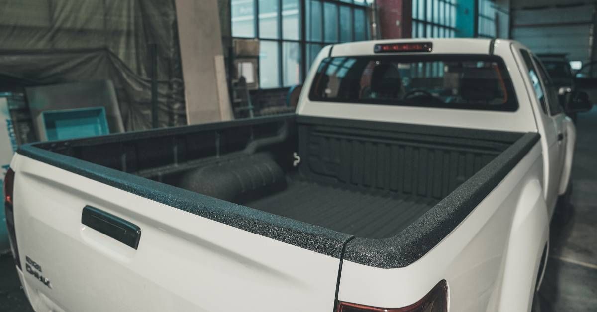 A white truck with a gray truck bed is parked in a large building. Another white truck is in front of the first one.