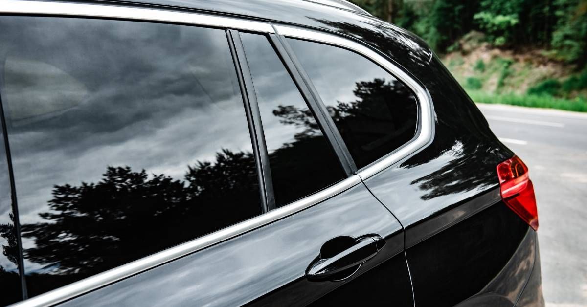A sleek black car with its rear windows tinted is parked outside next to the road and some trees during the way.