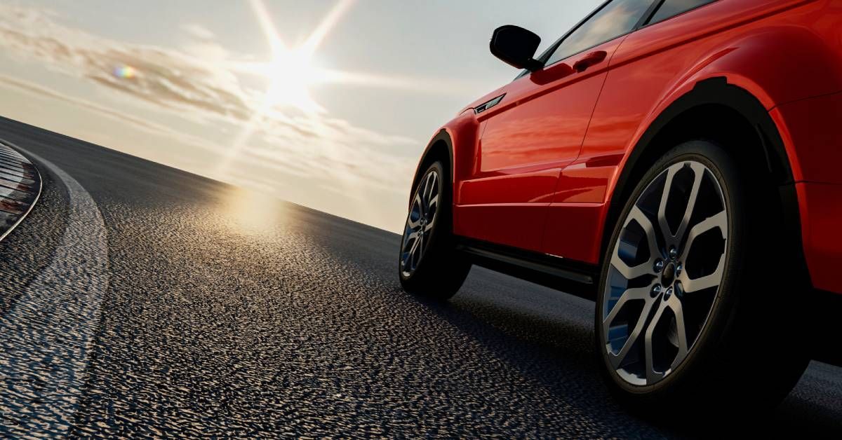 A shiny red car is driving on the road while the sun shines down from above, reflecting off the car’s exterior.