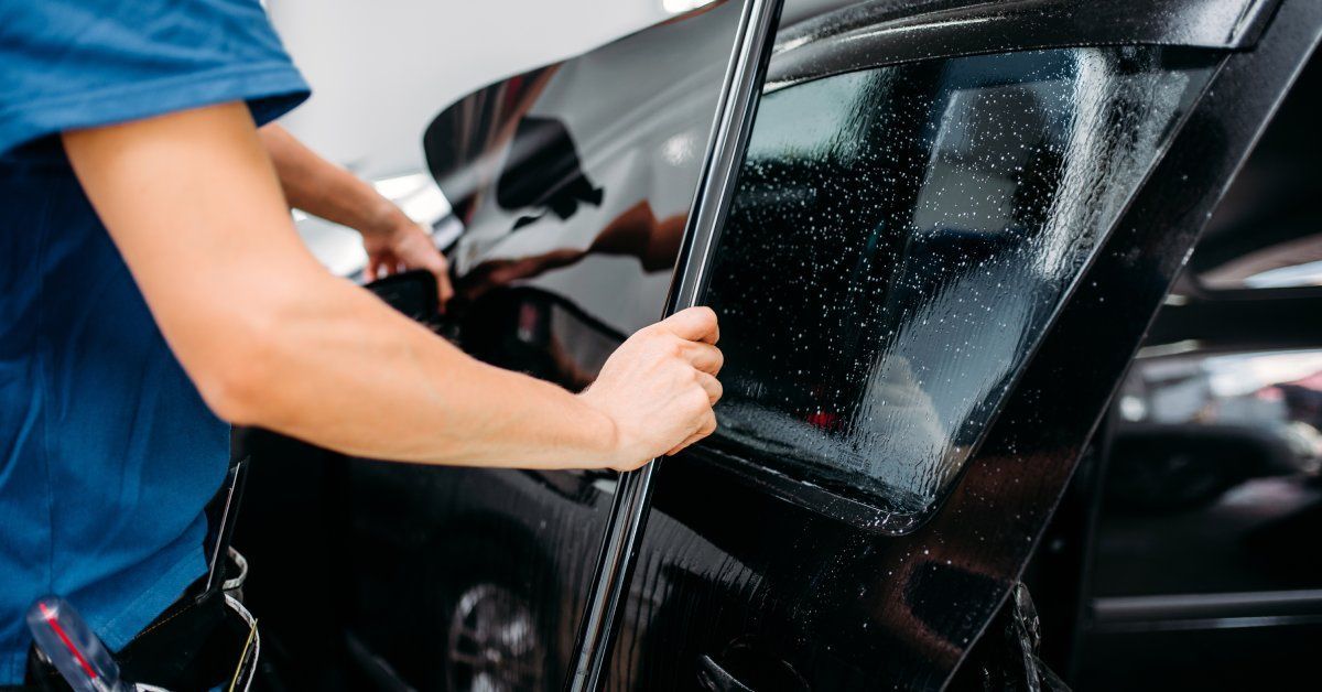 A person wearing a blue short-sleeved shirt is applying a window tint to a black car in a brightly lit space.