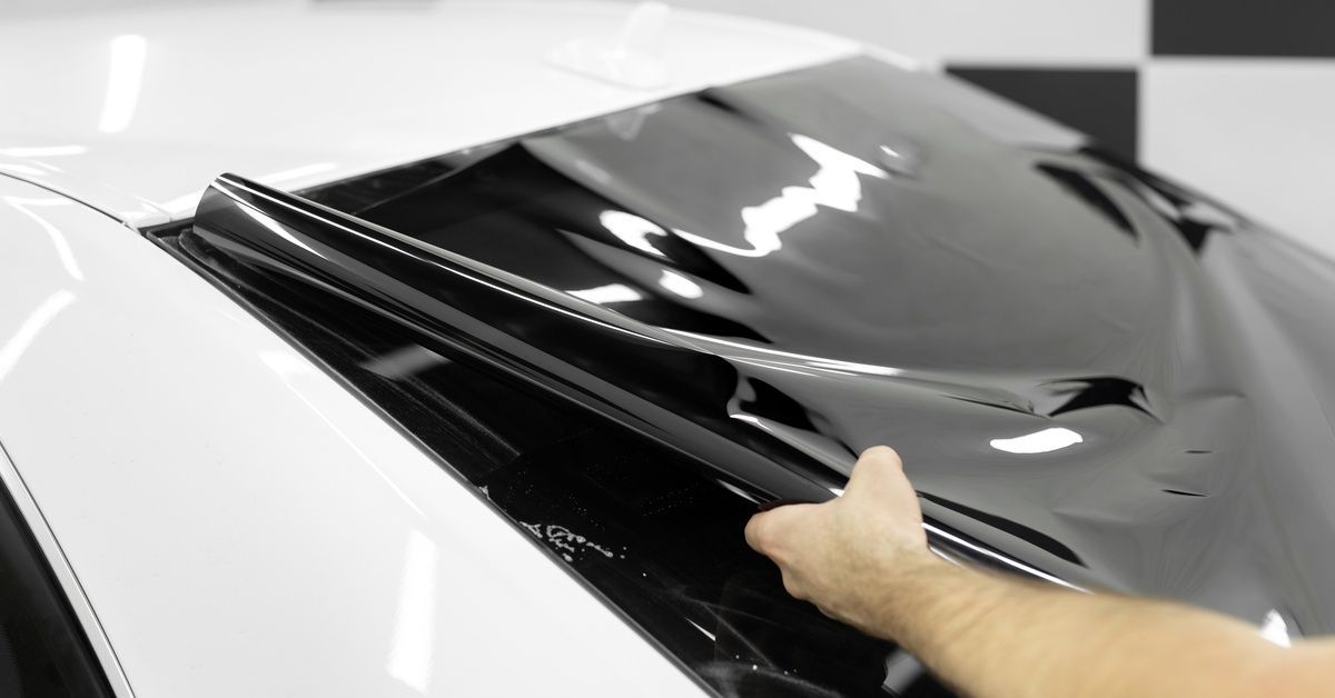 A person laying a large, sleek piece of black window tint film on the back window of a clean white car.
