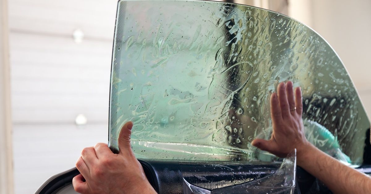 A person is placing a small tint film on a car window. The window tint appears wet as a hand flattens it out.