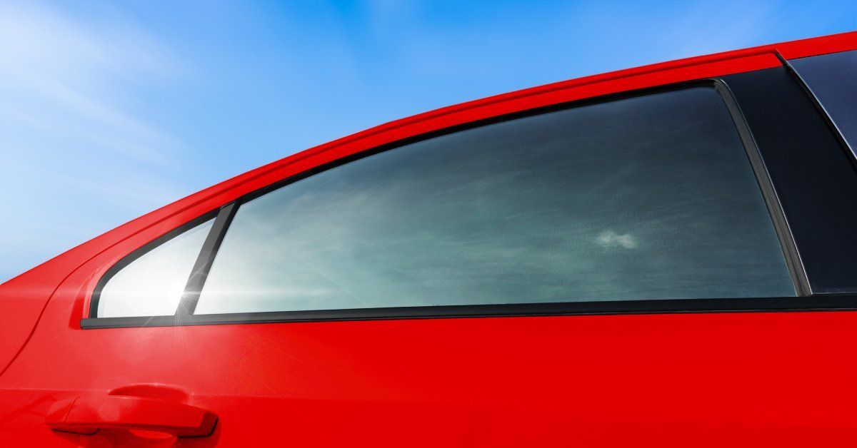 A red car parked outside on a sunny day with blue skies above. The car windows are tinted and reflecting the sky.