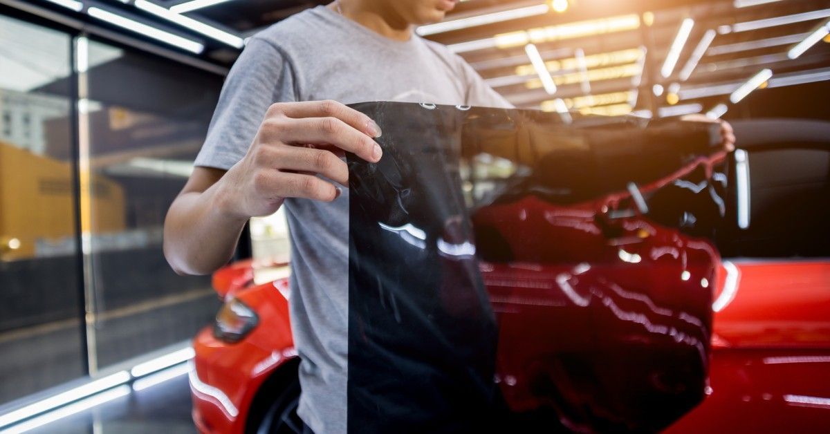 A person holding a large dark car tint film in their bare hands and standing in front of a bright re