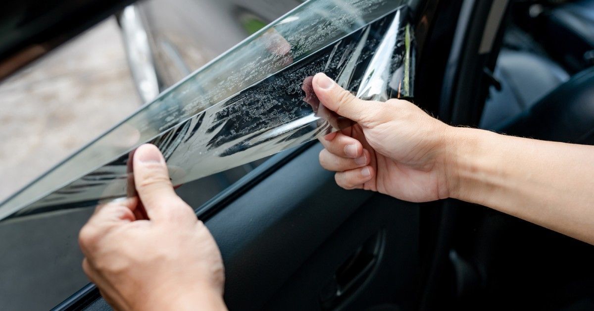 A person applying a window tint film on a car outside. The tint is being applied to the rear driver’s side window.