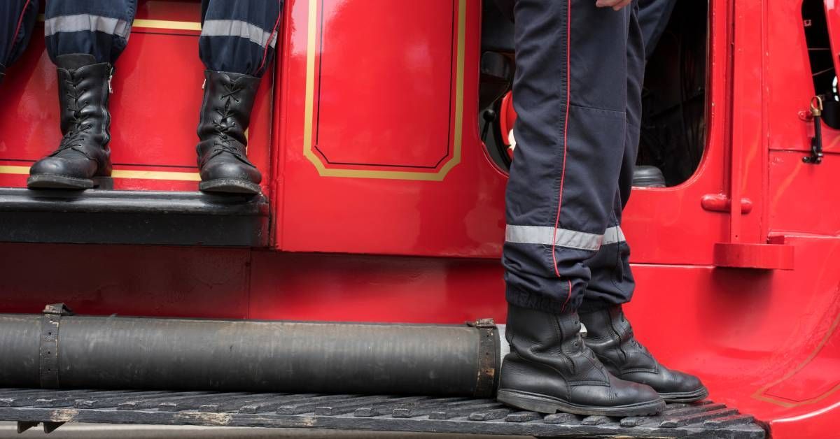 Three firefighters in black boots waiting on the side of a fire truck. One firefighter stands and the other two sit.
