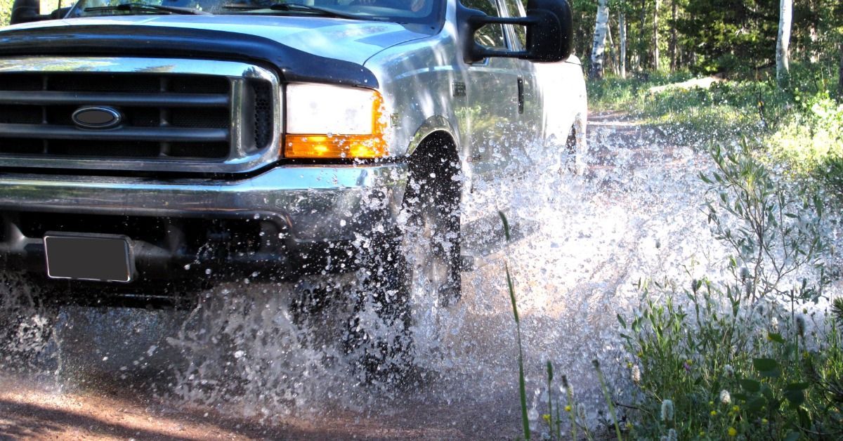 A person driving a white pickup truck through a large puddle on an off-road trail during the daytime.