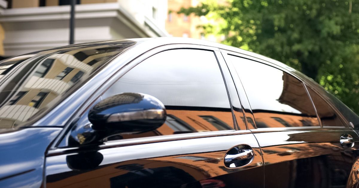 A black car with tinted windows parked outside during the daytime. The car sits in front of a large tree.