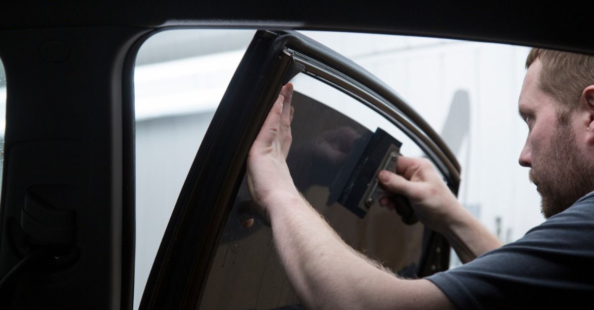 A professional using a small hand tool to apply a tinting film to the rear passenger’s side window o