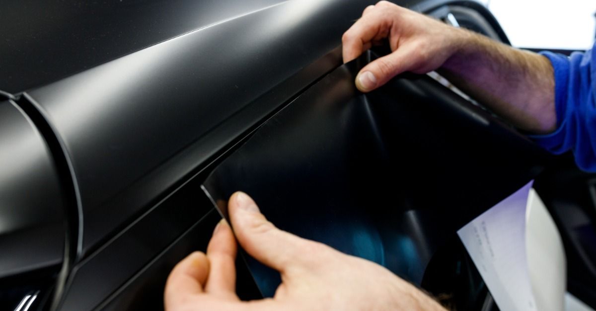A person wearing a blue shirt carefully applying a film tint with a matte black finish to the surface of a car window.