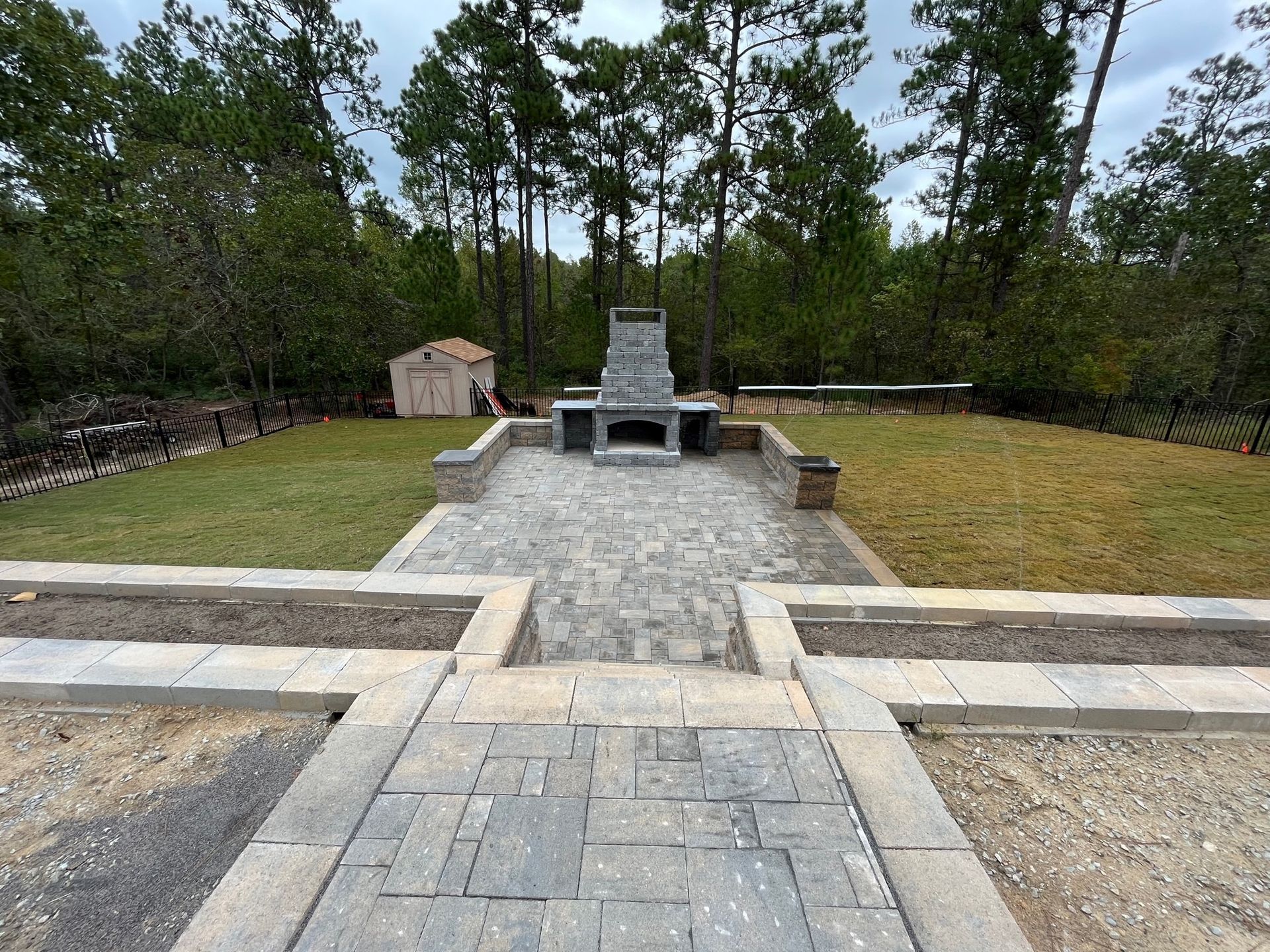 A brick walkway leading to a fireplace in a backyard surrounded by trees.
