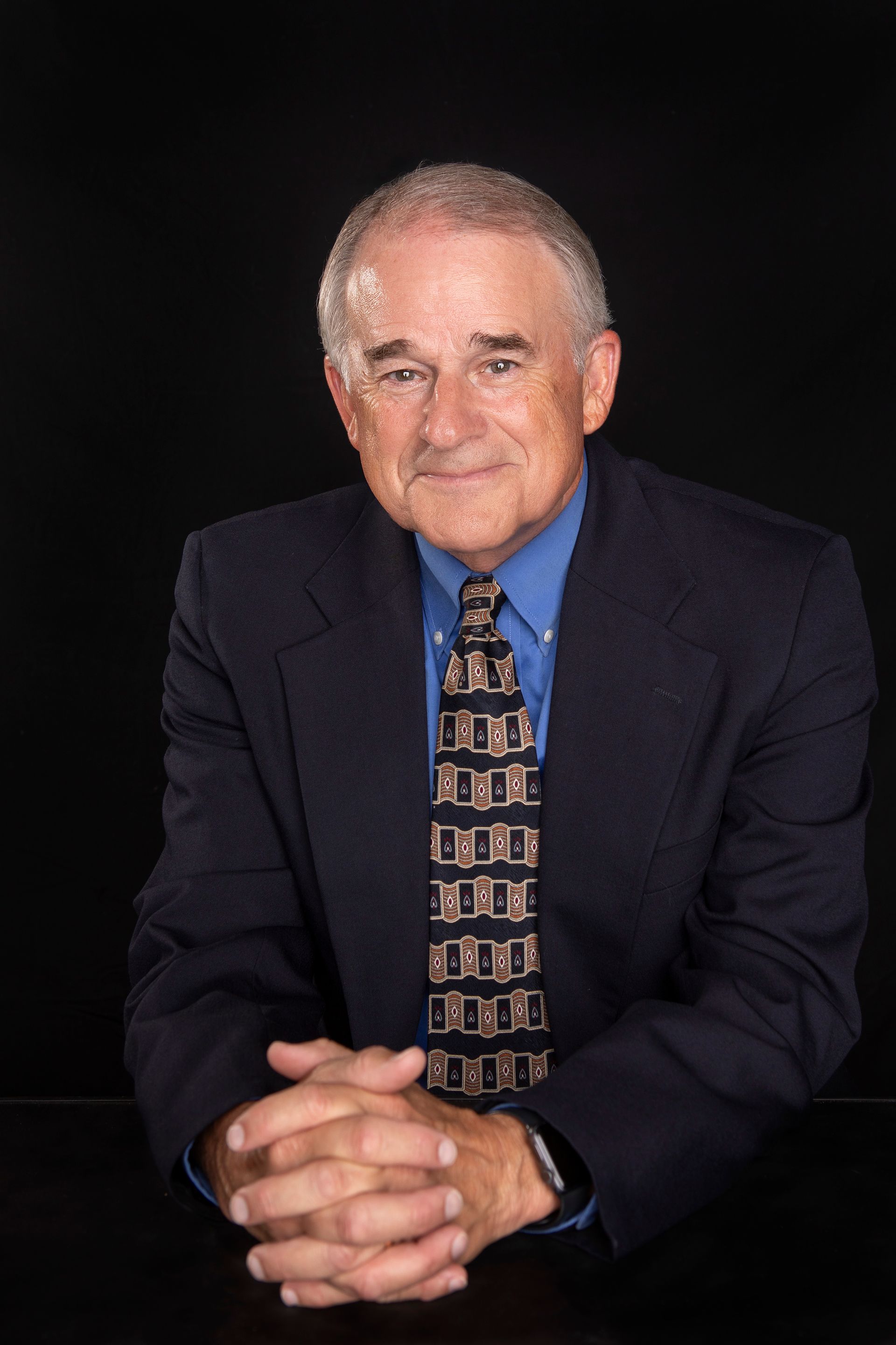 Man in a blue shirt and suit, hands clasped, smiling, seated against a black background.