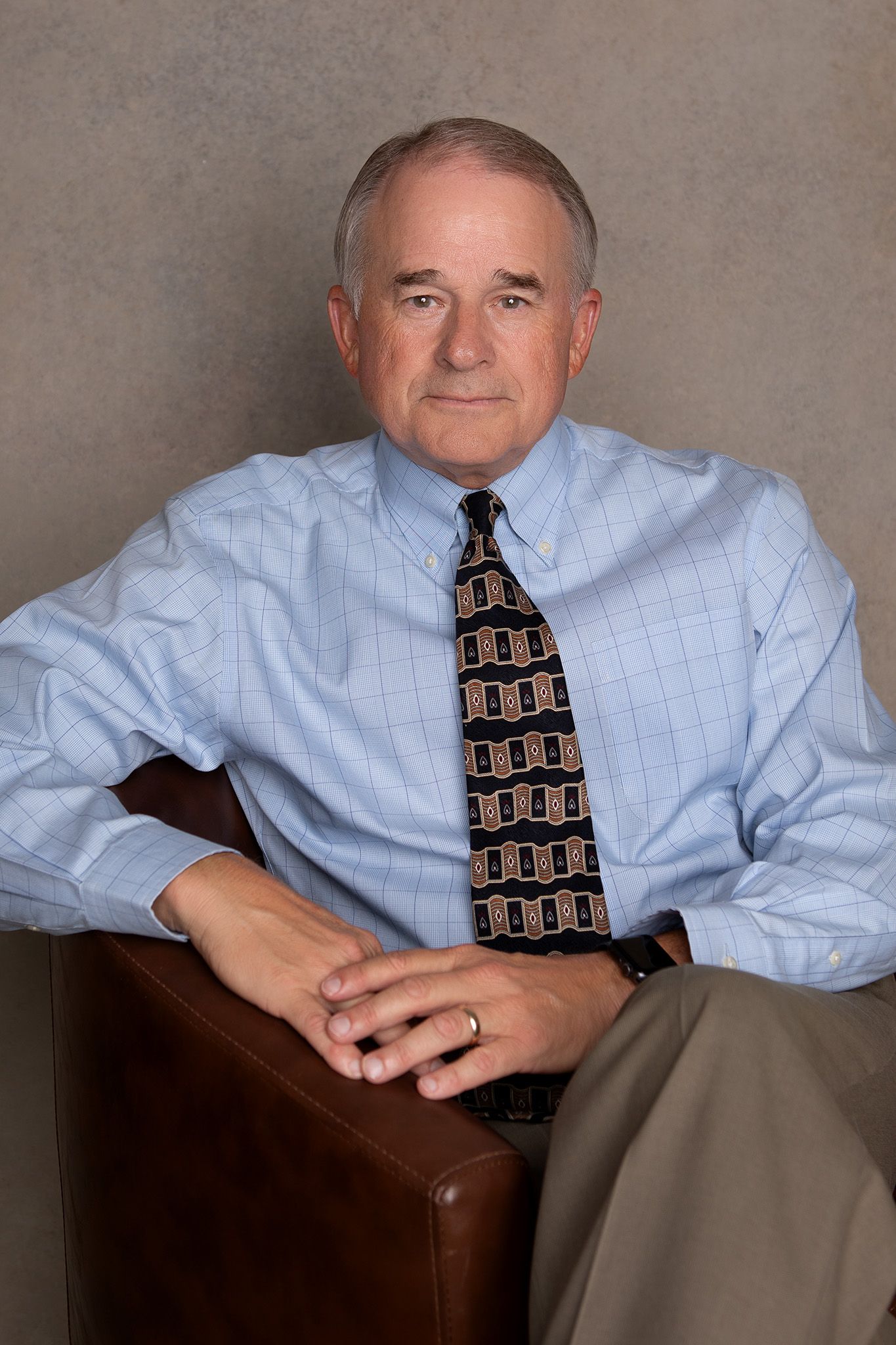 Older man in a blue shirt and patterned tie sits in a brown chair, arms crossed.