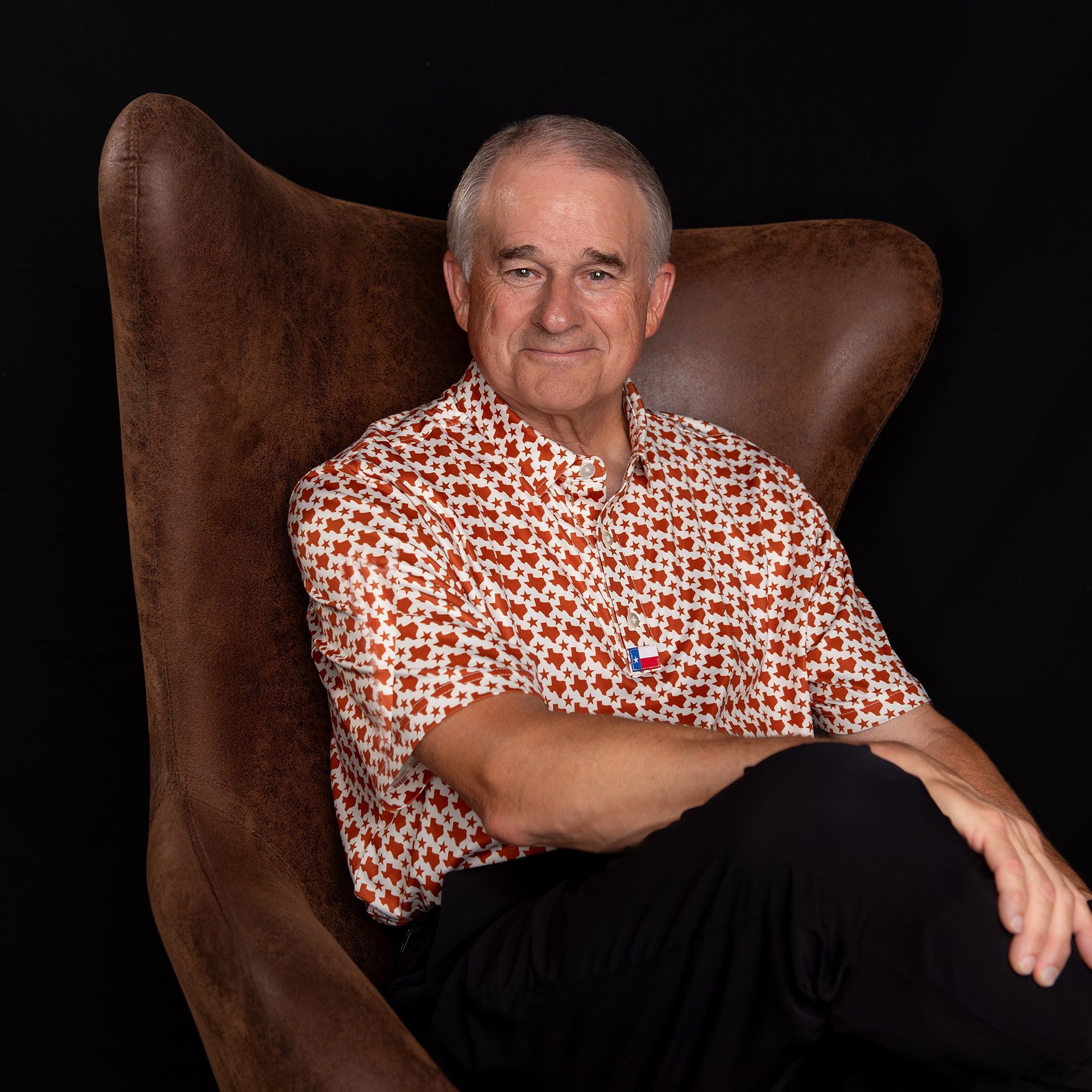 Man in patterned shirt sits in a brown leather armchair, smiling against a black background.