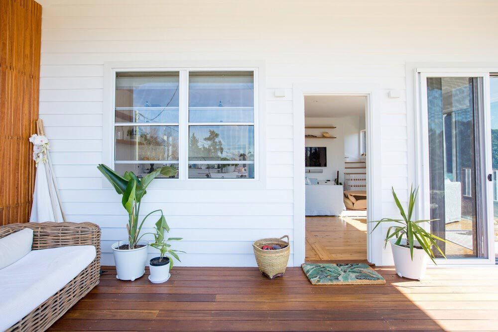 A Wooden Deck With a Couch and Potted Plants in Front of a White House — DeBu Studios In Russell Vale, NSW
