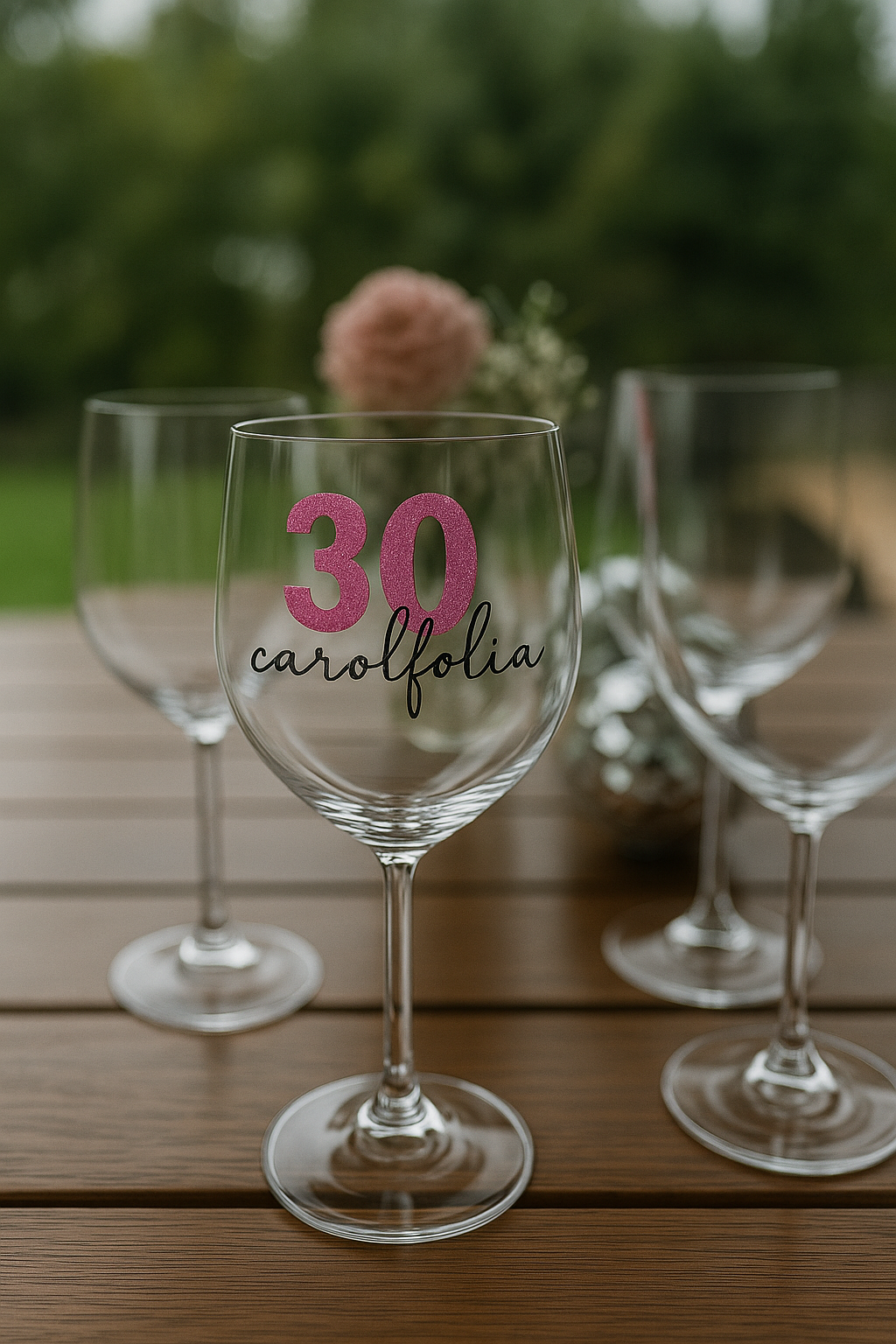 Taças de vinho sobre uma mesa de madeira, uma delas com a inscrição “30” e “linda”, com um arranjo de flores rosa ao fundo.