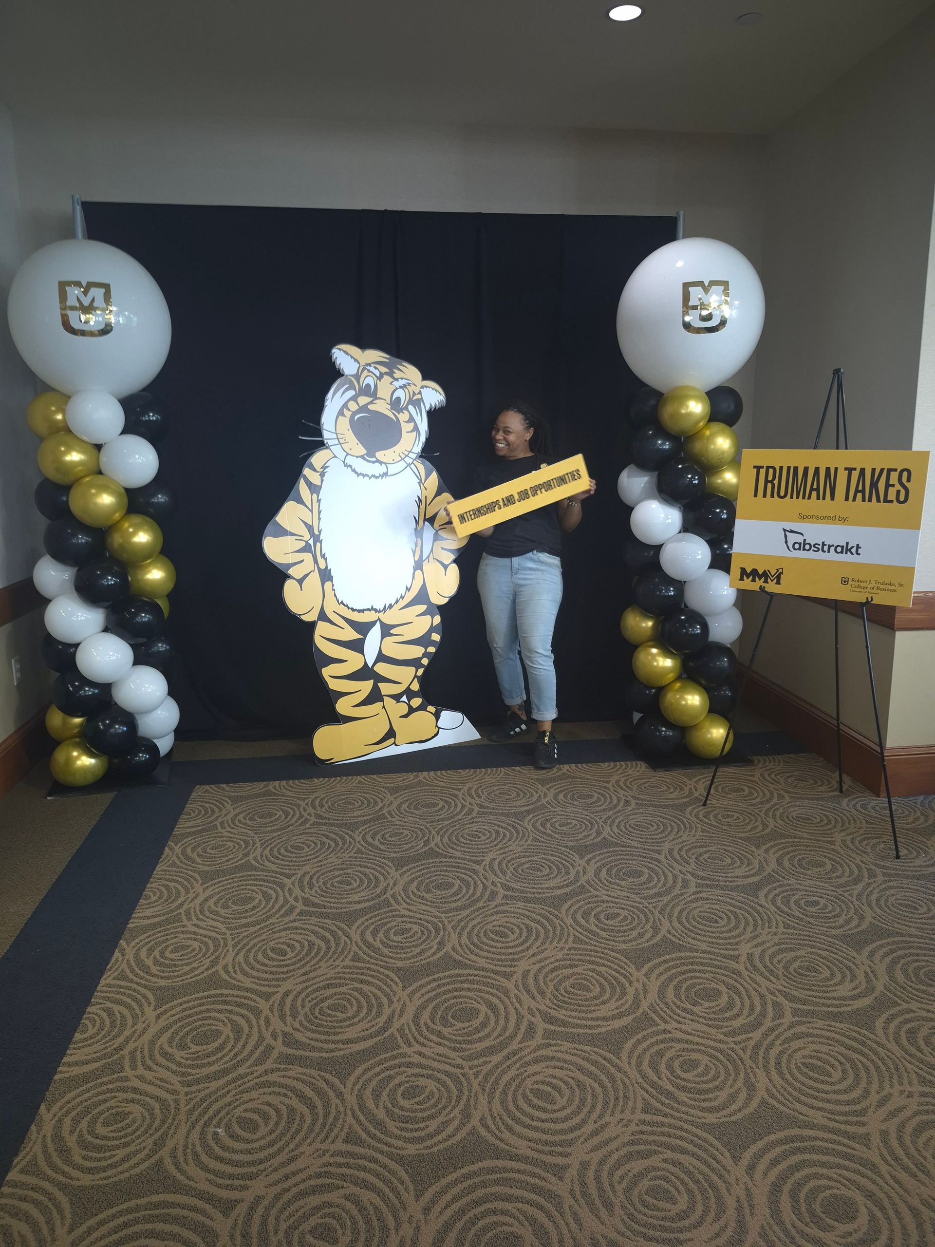 Person posing beside a lit tiger cutout between black, white, and gold balloon columns at an indoor event.