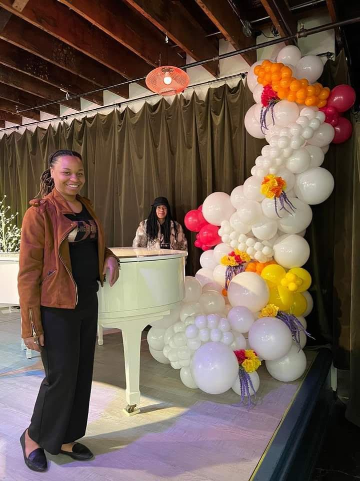 Woman on a stage beside a white piano and a balloon arch, smiling at an indoor event.