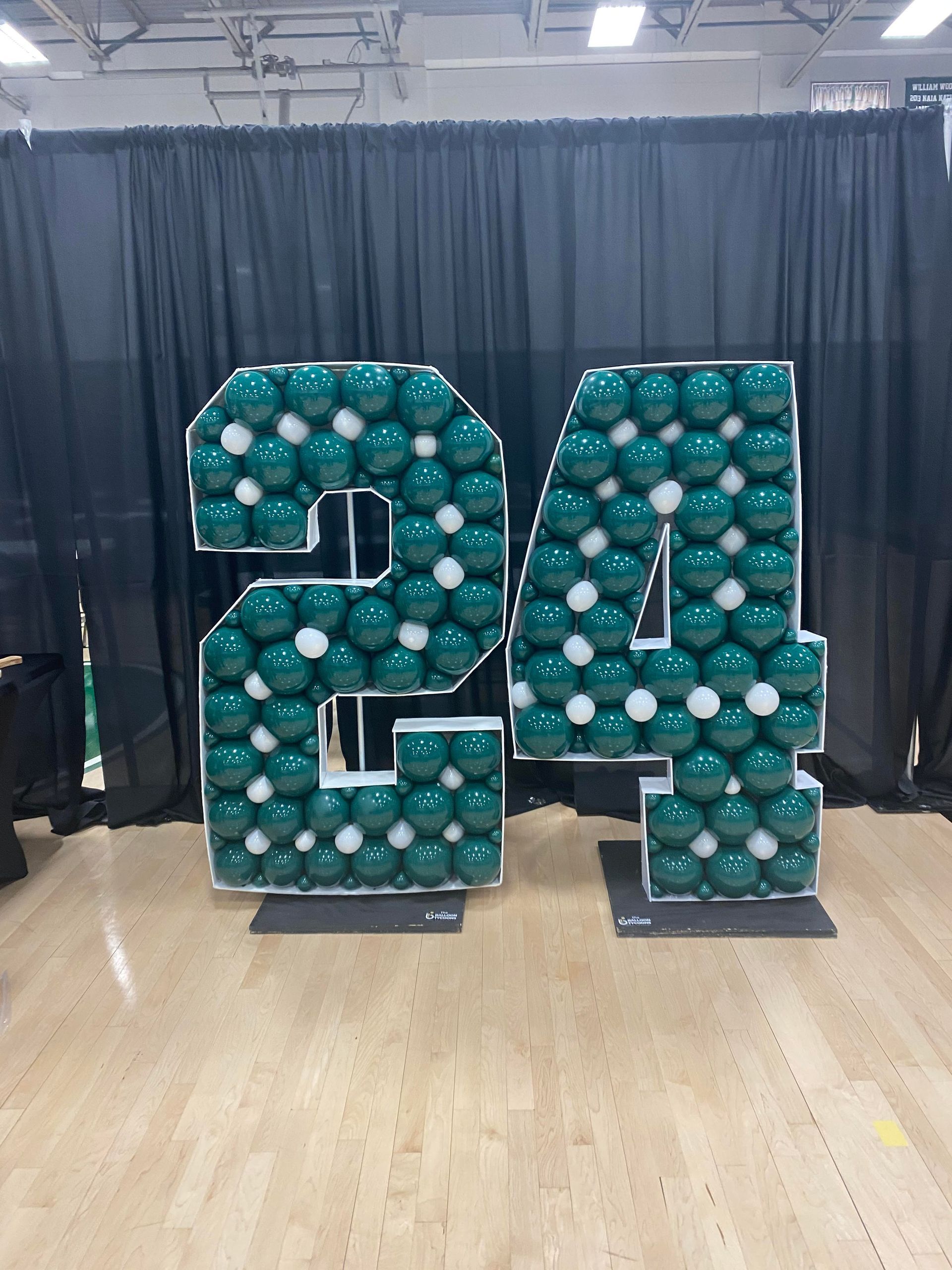Teal balloon numbers “24” on a stage with black curtains and wooden floor