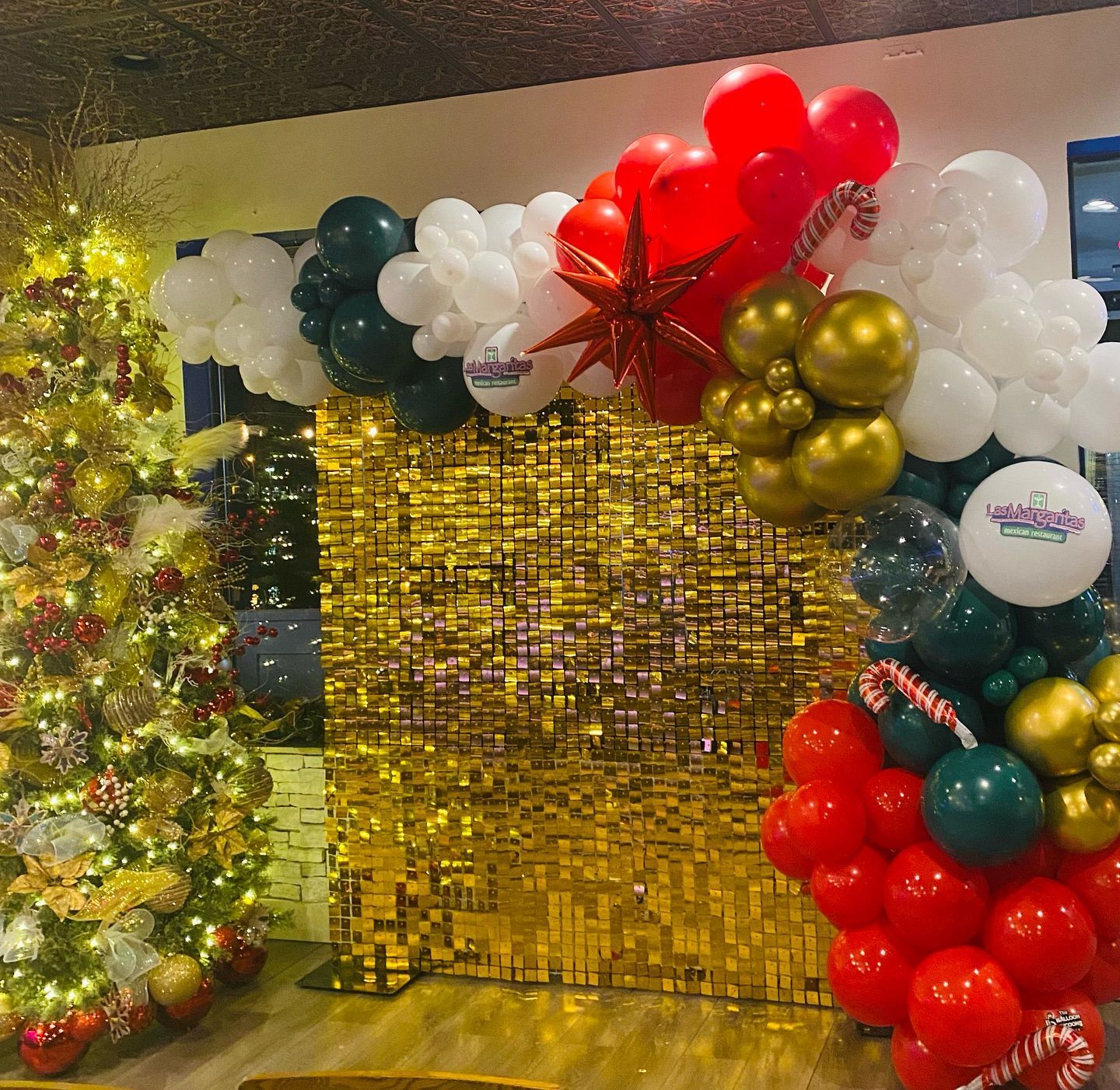 Gold sequin backdrop with red, white, teal, and gold balloon arch in a decorated event space
