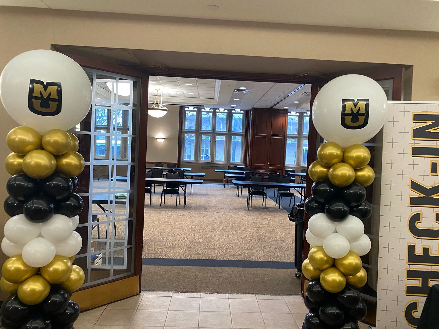Mizzou balloon arch with black, gold, and white balloons framing open doors to an indoor lobby