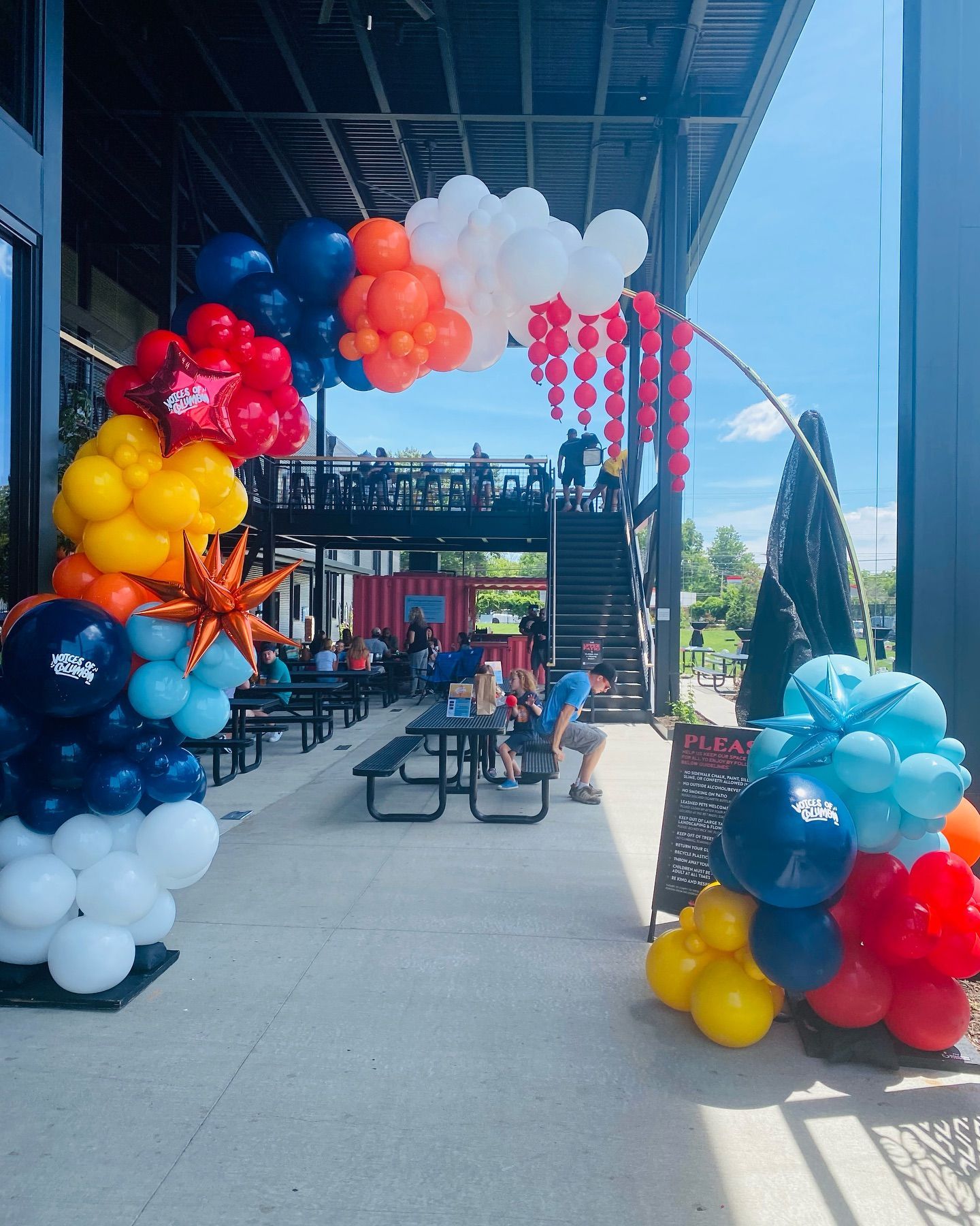 Colorful balloon arch and clusters frame an outdoor patio walkway with tables and chairs under a covered roof.