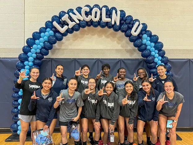 Lincoln-U volleyball team posing under blue balloon arch in a gym.