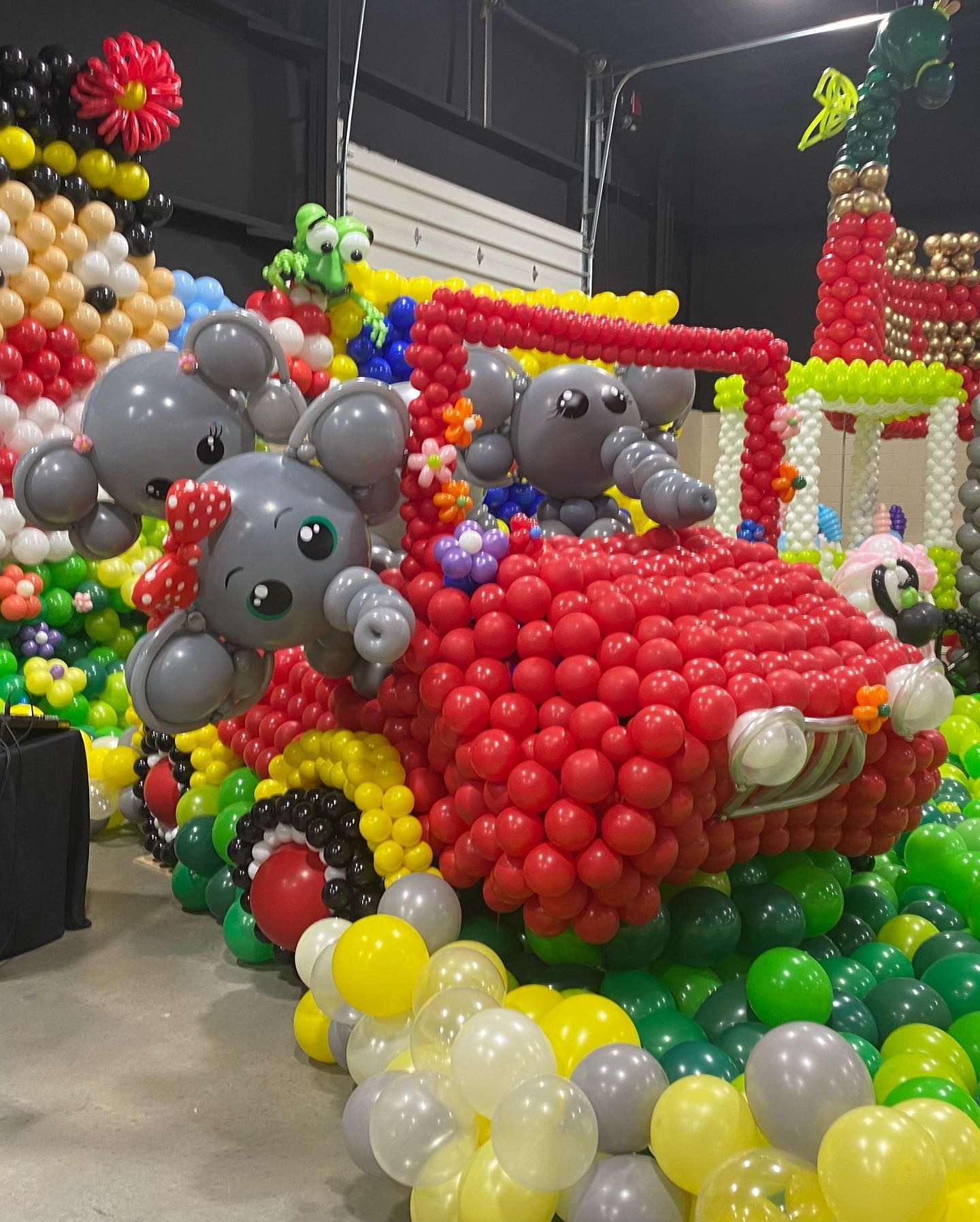 Colorful balloon sculpture of gray elephants riding in a red car at an indoor event