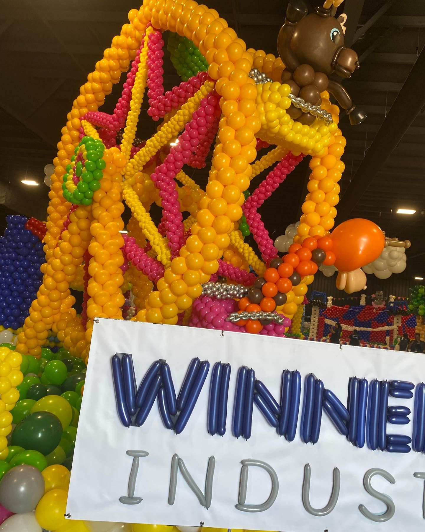 Balloon sculpture display with a white “WINNIE” sign in a festival-like indoor setting