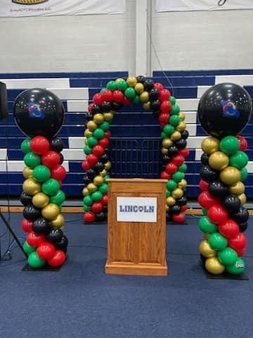 Balloon arch and balloon columns framing a podium with a Lincoln sign in a gymnasium