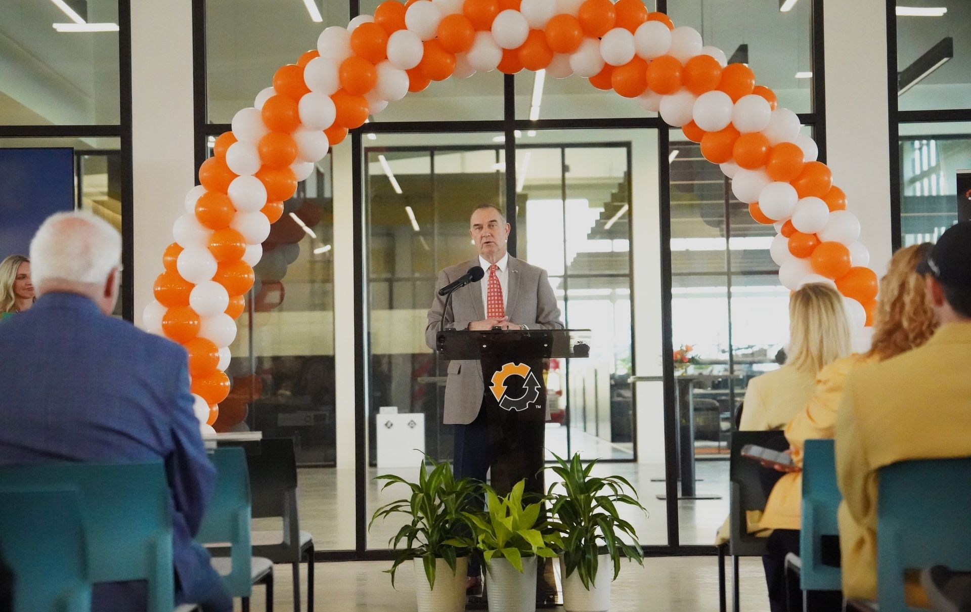 Speaker at podium under orange-and-white balloon arch, addressing seated audience indoors