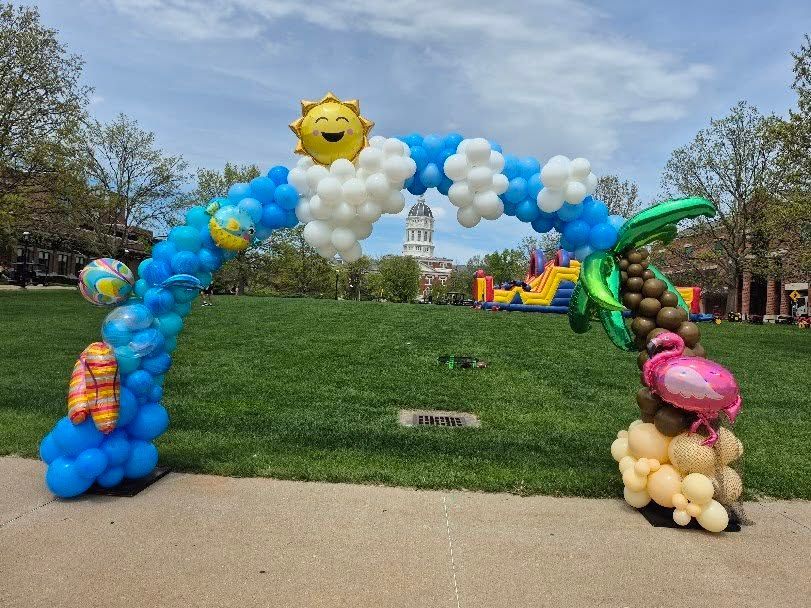 Balloon arch with smiling sun over a park path beside a grassy hill and trees