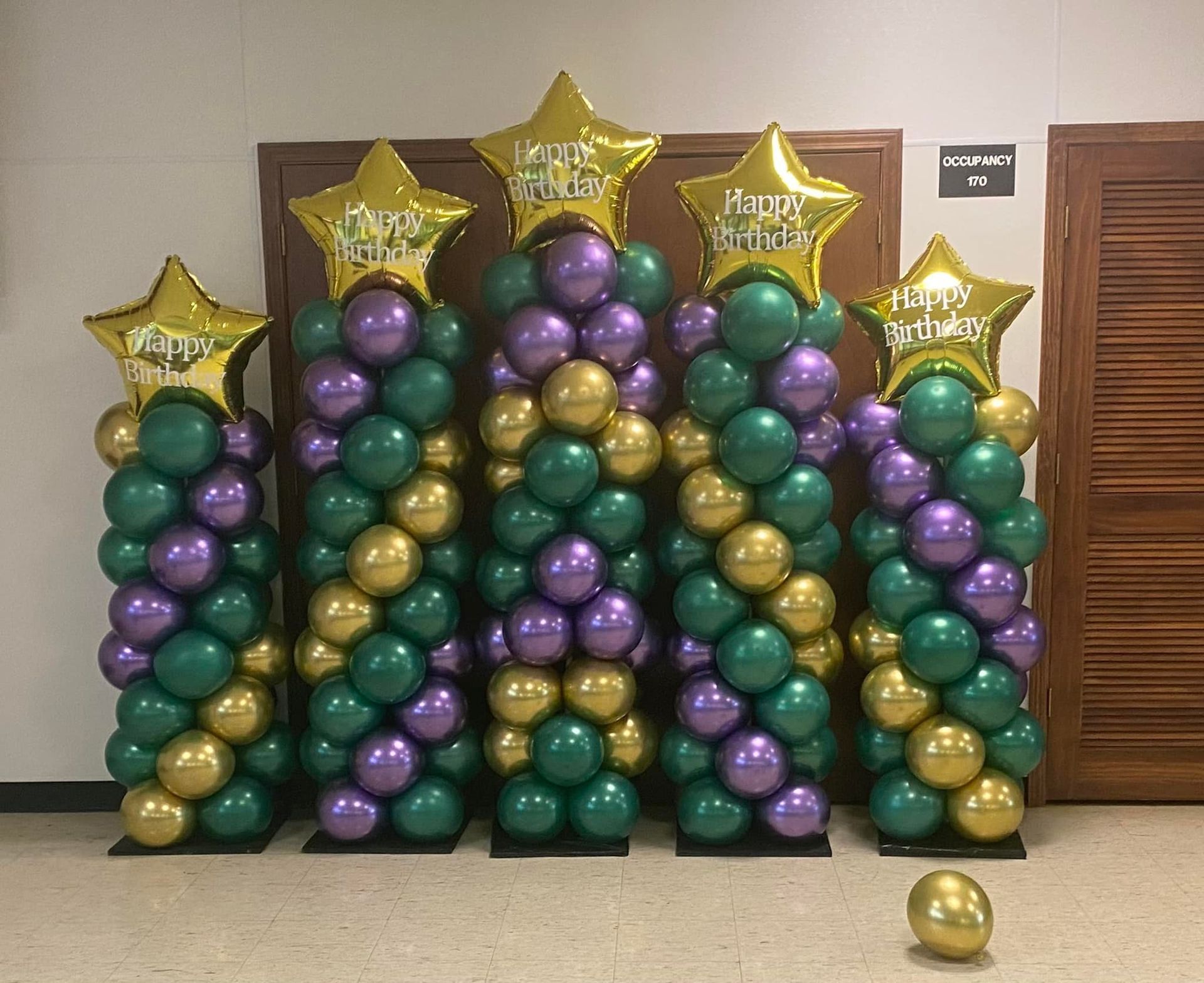 Balloon display of green, purple, and gold stars and columns against a wall, with one loose gold balloon on the floor