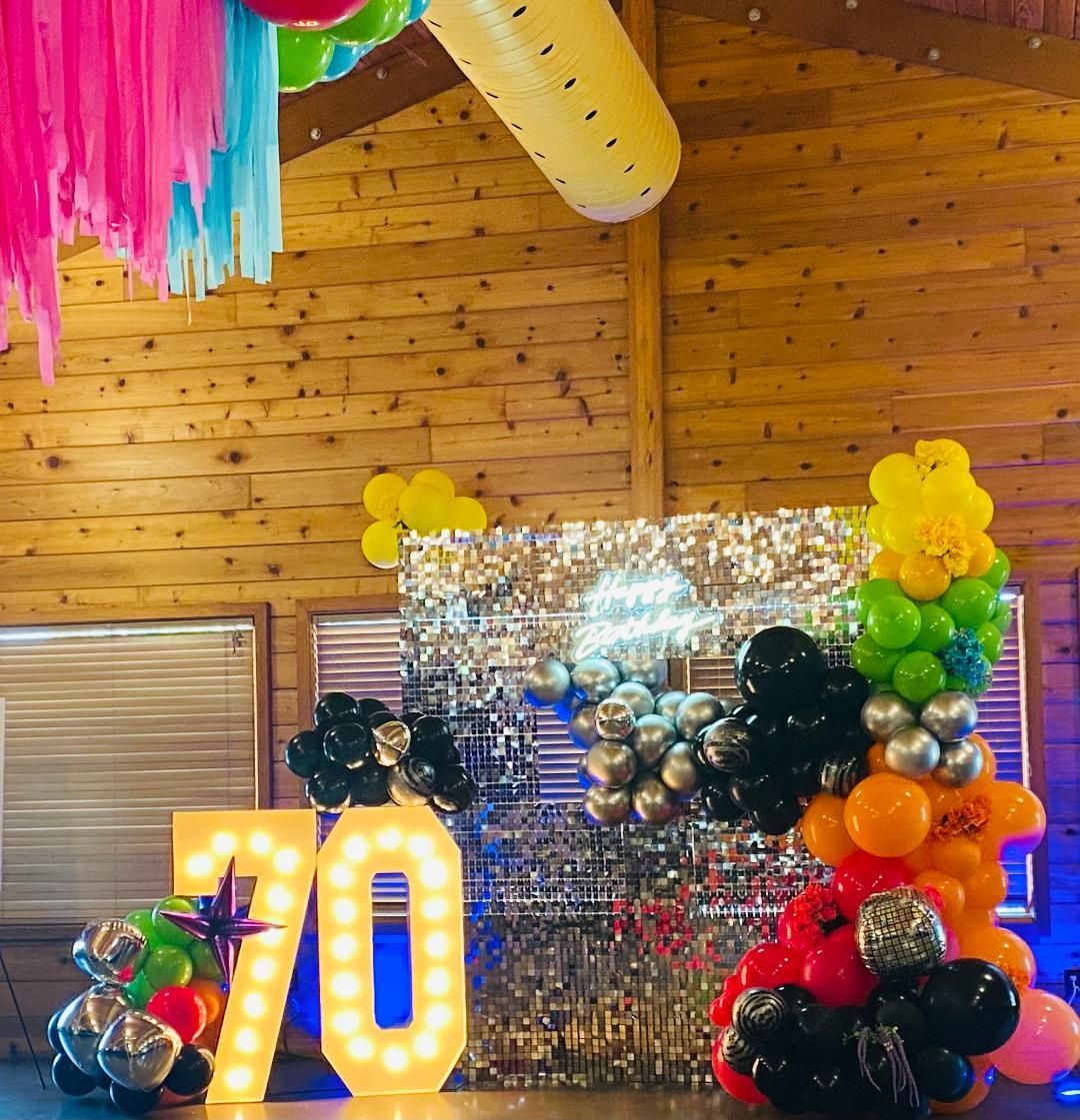 Birthday party backdrop with glowing “70” numbers, balloons, and silver sequin decorations in a wood-paneled room
