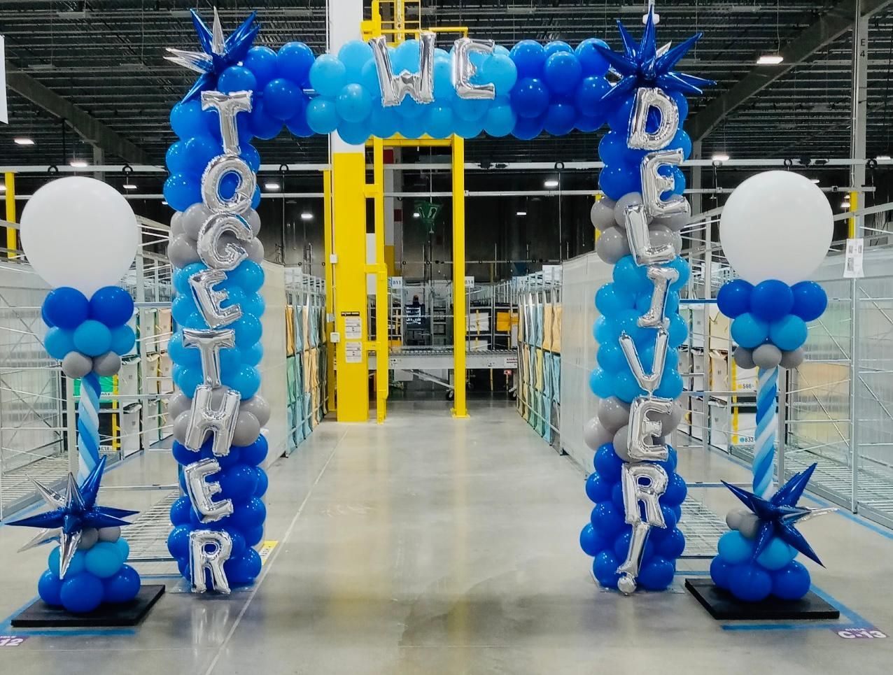 Blue balloon arch with white “Welcome” letters at a store entrance, flanked by balloon columns and stars.