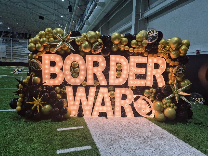 Illuminated “BORDER WAR” sign with gold balloon decorations on a sports field, against a dark background
