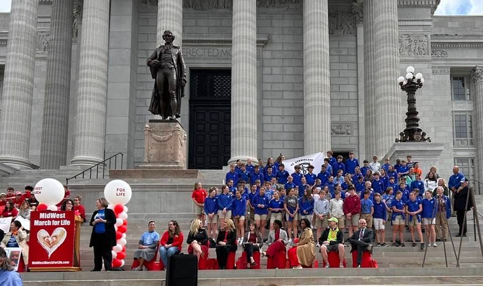 Large group in blue and red outfits posed on courthouse steps with statues and columns behind them