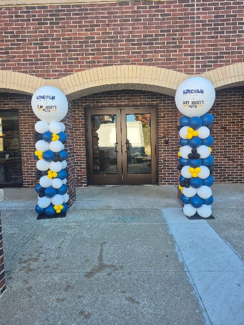Balloon columns with blue, white, and yellow balloons framing a brick building entrance.