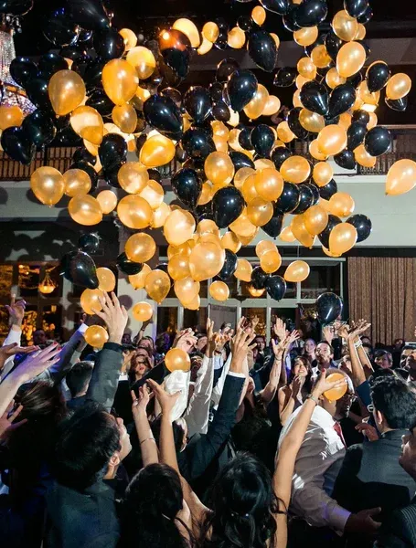 Crowd reaching up as black and orange balloons fill the air at an indoor celebration