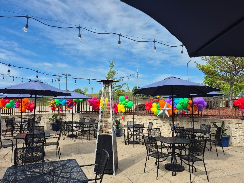 Outdoor patio with string lights, black tables, umbrellas, and colorful balloons by a stone wall