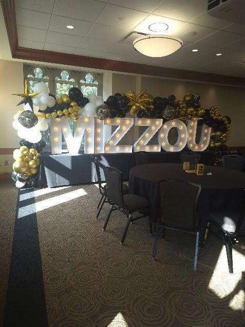 Decorated event room with large “MIZZOU” letters, black chairs, tables, and gold-and-white balloons