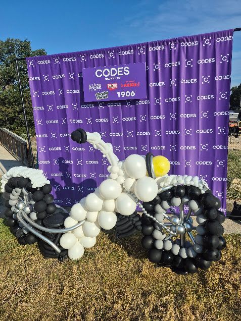 Balloon sculpture of a bicycle in front of a purple CODES backdrop outdoors