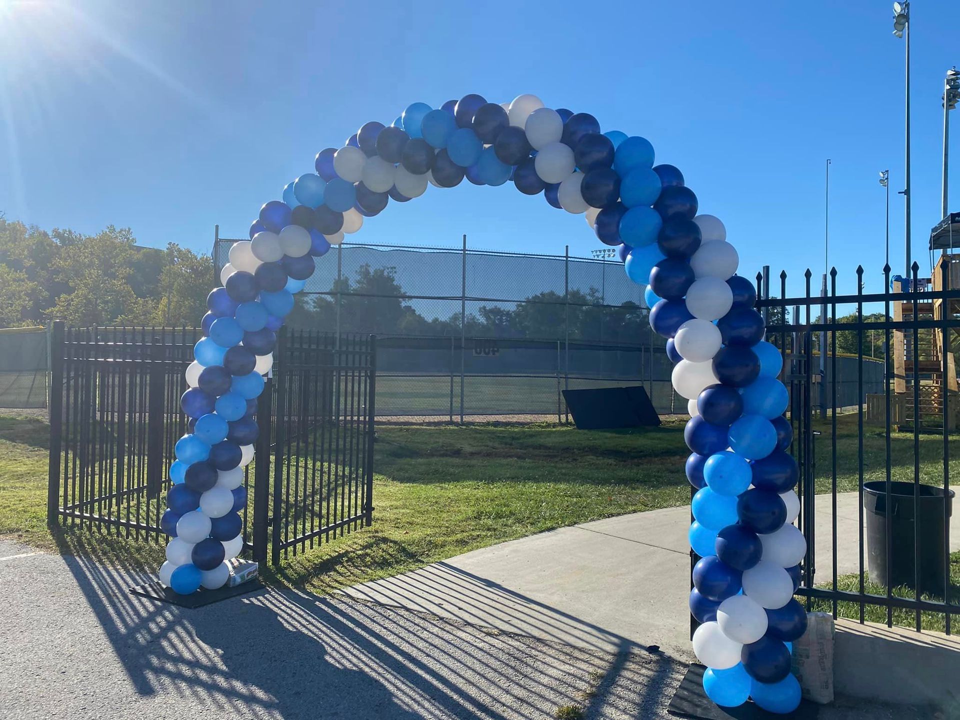 Blue and white balloon arch at a sports field entrance on a sunny day