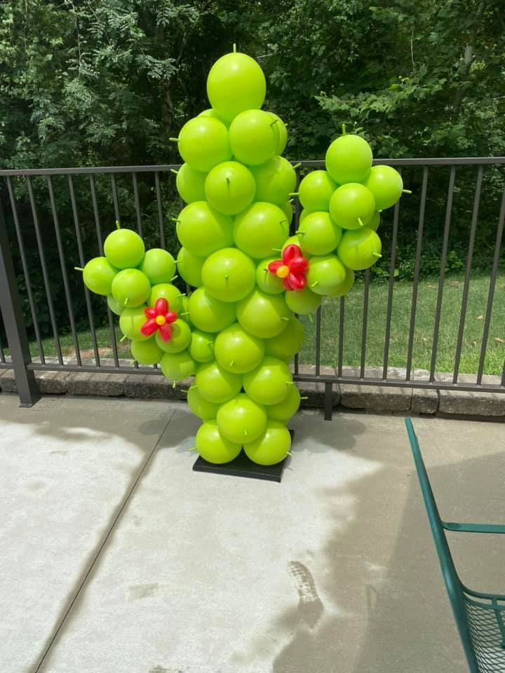 Green balloon cactus sculpture outdoors on a patio, with small red balloon flowers and a black railing behind it