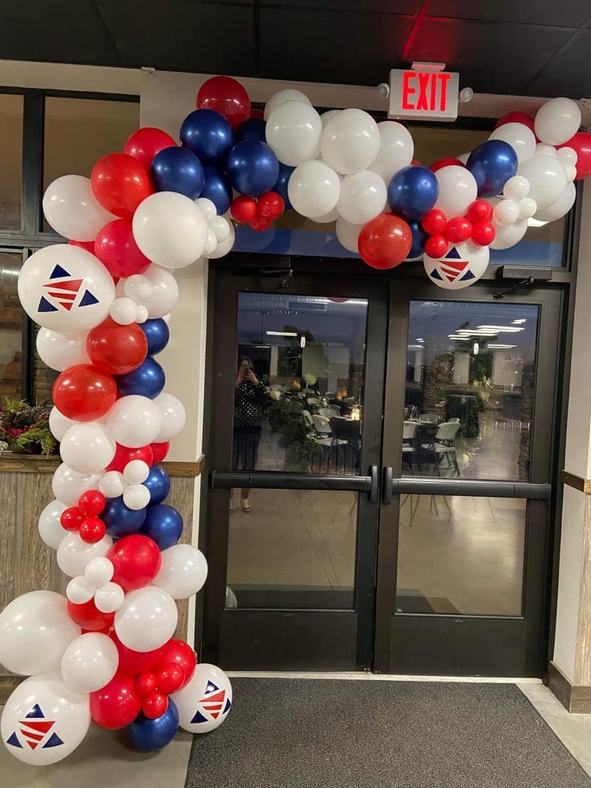 Red, white, and blue balloon arch framing glass double doors with an exit sign overhead.