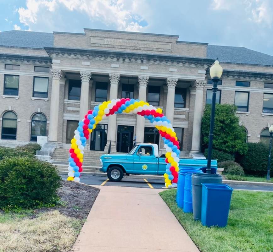 Vintage blue pickup under rainbow balloon arch in front of a stone building