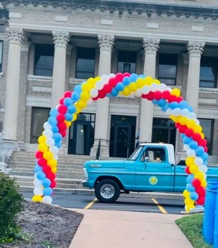 Blue vintage pickup truck under a red, yellow, white, and blue balloon arch in front of a building