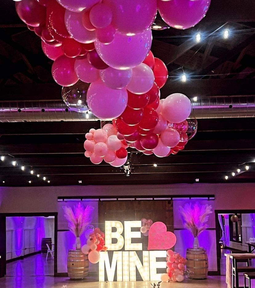 Pink and white balloon arch above illuminated “BE MINE” sign in a decorated event hall