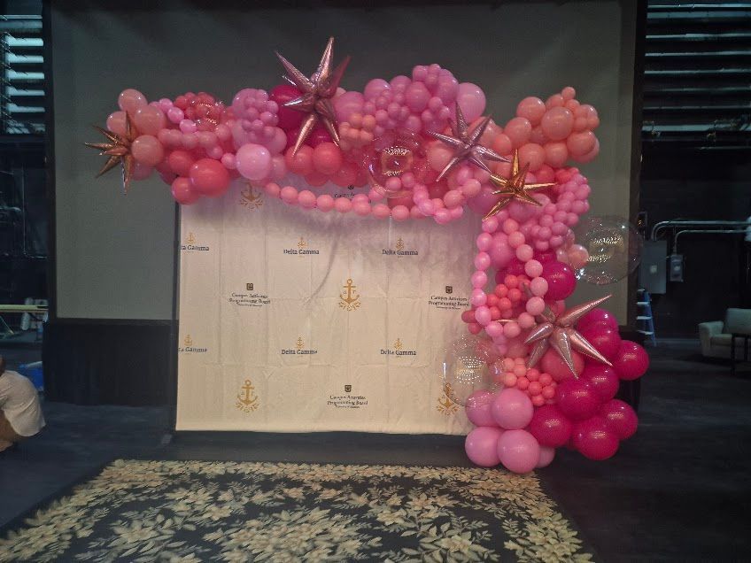 Pink balloon arch around a beige step-and-repeat backdrop in a dimly lit event space.